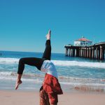 woman wearing red jacket and black leggings doing hand stand in seashore